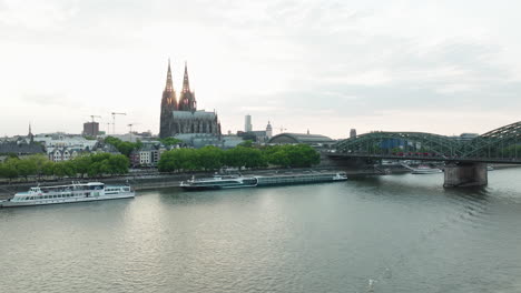 Drohne-Filmt-Ein-Schiff-Auf-Dem-Rhein-Herein,-Die-Drohne-Fährt-Hoch-Und-Man-Sieht-Den-Kölner-Dom-Im-Sonnenuntergang,-Ein-Zug-Fährt-Auf-Der-Hohenzollerbrücke-über-Den-Rhein