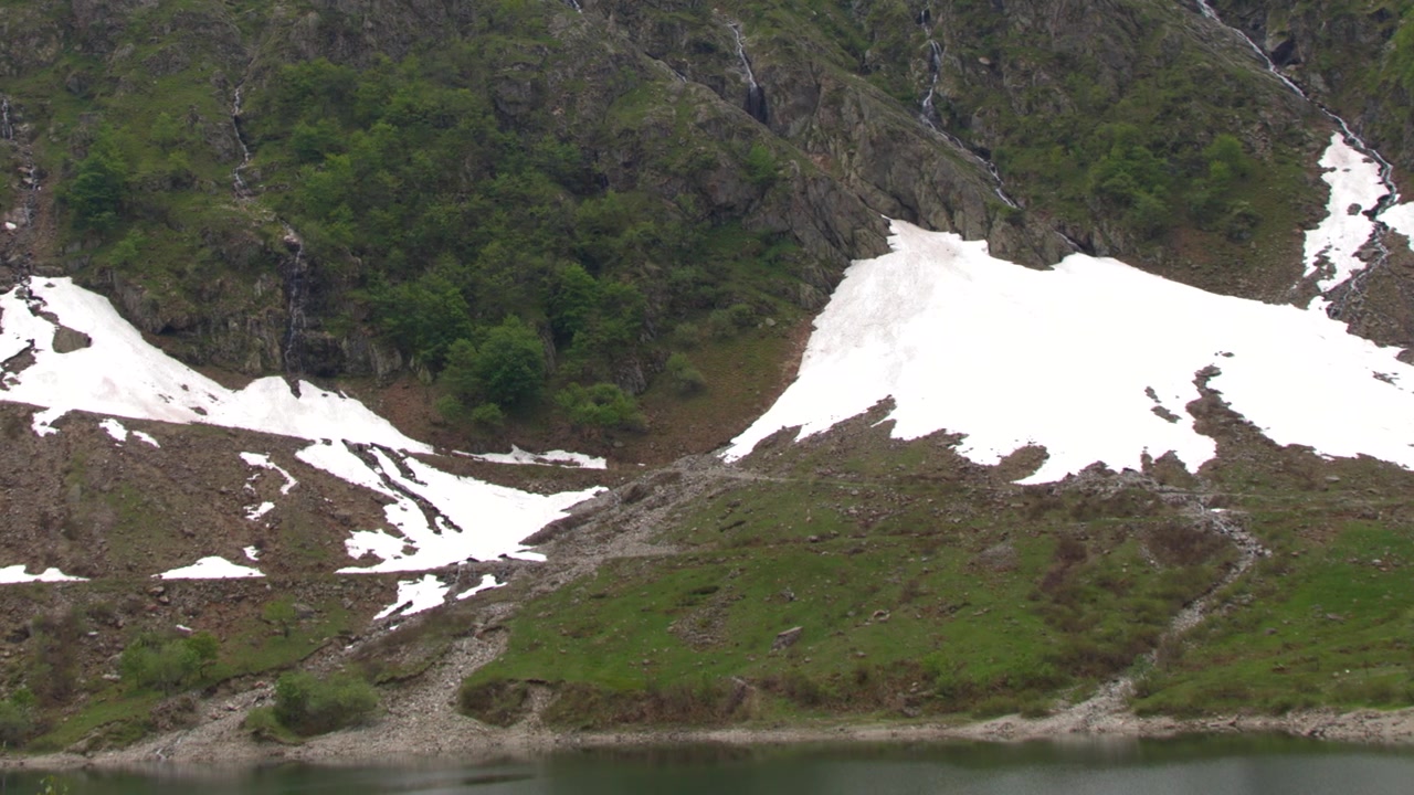 Statisk vidvinkelbilde av smeltende snø på steinete fjellsider over en rolig innsjø