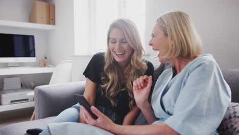 senior mother with adult daughter sitting on sofa looking at pictures on mobile phone