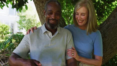 Diverse-senior-couple-in-garden-taking-selfie-sitting-under-a-tree-and-smiling