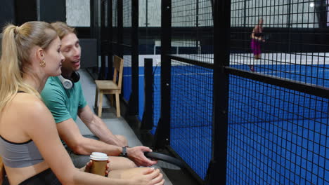 man and woman enjoying padel tennis match, with rackets and coffee, watching game, on indoor court