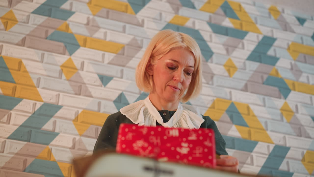 Donna anziana che apre un regalo rosso di Natale in un interno accogliente con parete piastrellata geometrica, che indossa un colletto di pizzo, che scarta delicatamente il regalo con un sorriso caldo e luci natalizie soffuse, primo piano intimo che cattura.