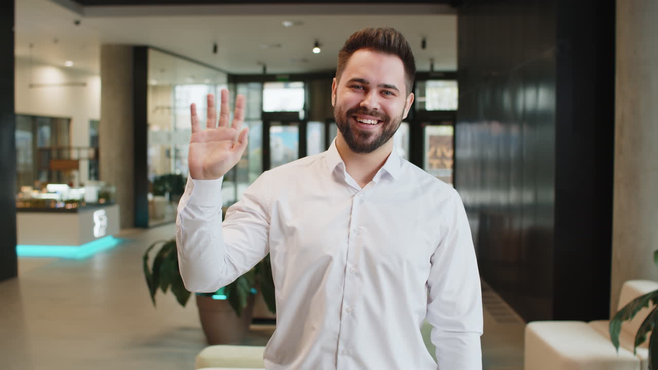 Happy young caucasian businessman looking at camera and waves hand hello in modern office lobby