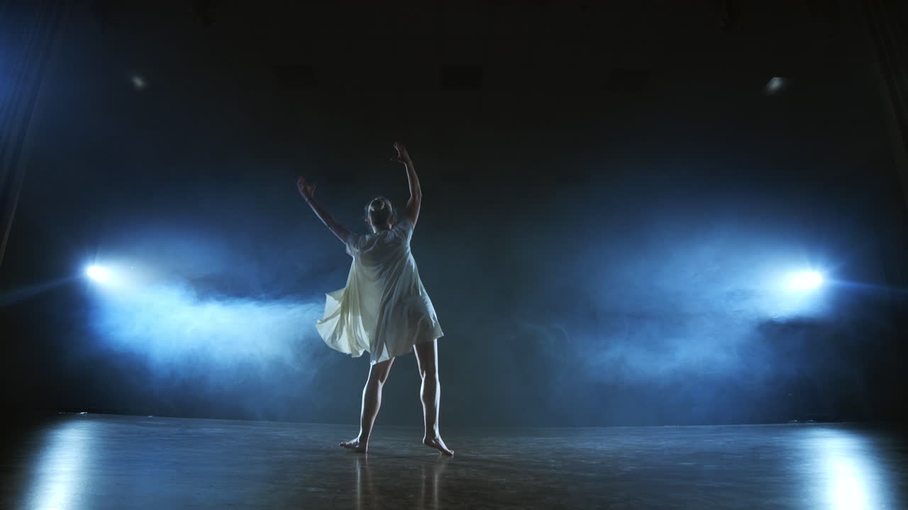 Dramatic spins and flips of a modern ballet dancer from a musical. A single woman emotionally dances on stage against a dark background with smoke in the spotlight in slow motion.