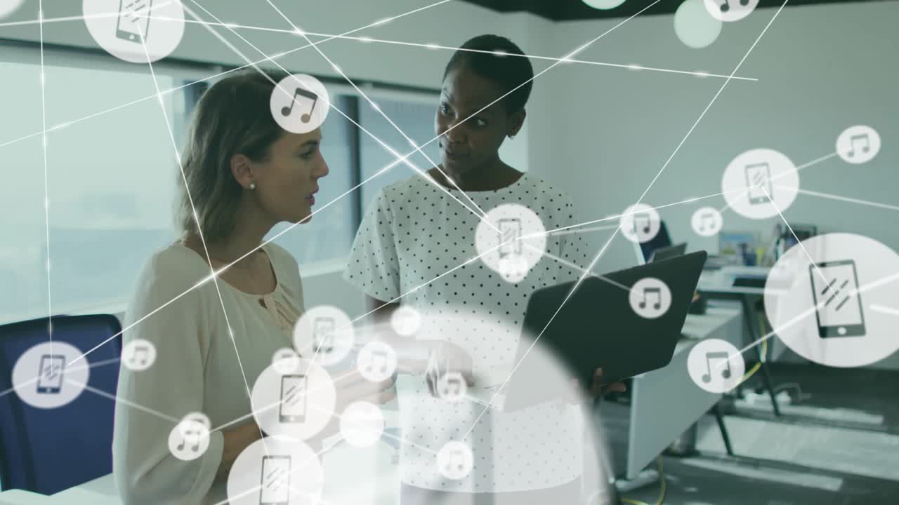 Two women collaborating over laptop in office, visualizing tech with floating phone and music icons