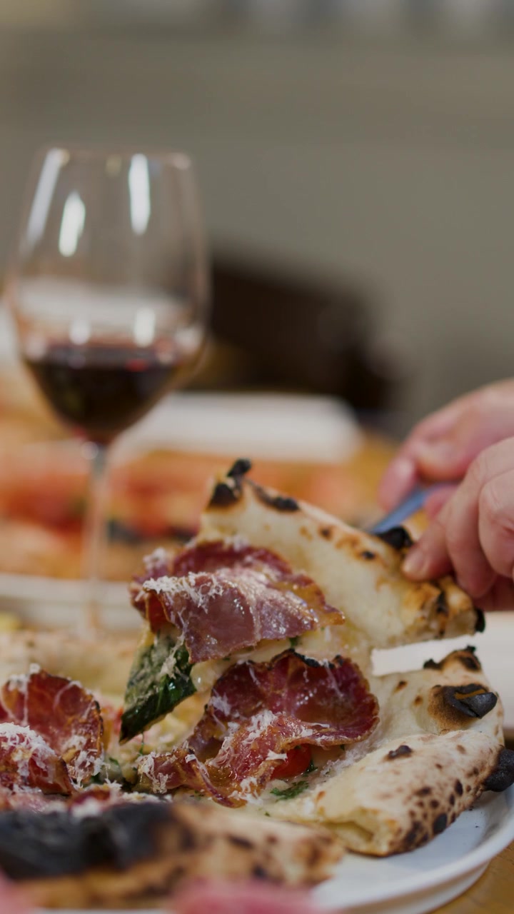 Primo piano di mani che usano una forchetta per mangiare pizza gourmet con prosciutto e vino rosso