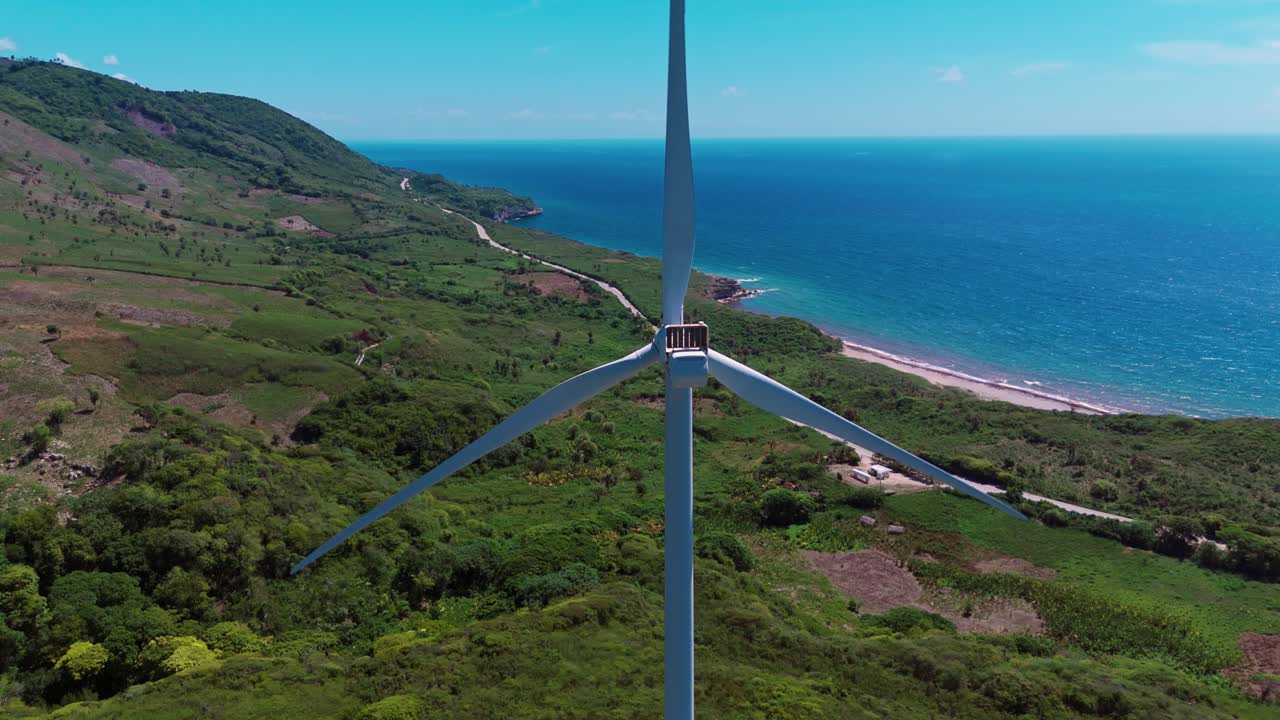 Close-up aerial drone shot of wind turbine blades spinning, generating clean energy with green hills and blue ocean in background, Dominican Republic