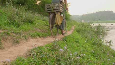 Toma-En-ángulo-Bajo-De-Un-Agricultor-En-Un-Ciclo-En-Un-Camino-Graval-A-Lo-Largo-De-La-Orilla-Del-Río-En-Loc-Binh-A-Lo-Largo-Del-Distrito-Rural-De-La-Provincia-De-Lạng-Sơn-En-La-Región-Noreste-De-Vietnam