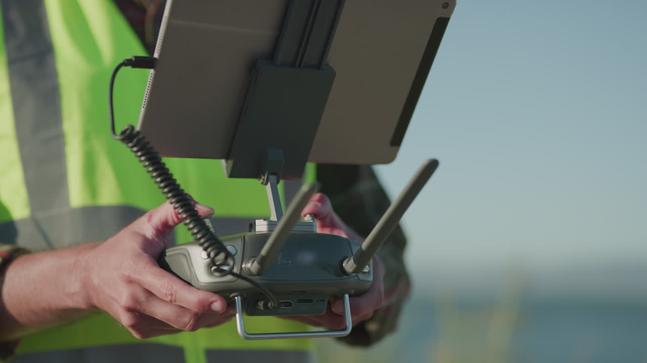 낮에는 안전 조끼를 입은 남자가 태블릿이 연결된 라디오 드론 컨트롤러를 들고 야외에 서 있습니다.