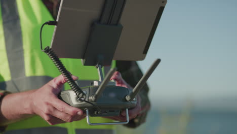 A-man-in-a-safety-vest-stands-outdoors-during-the-day-holding-a-radio-drone-controller-with-a-tablet-plugged-into-it