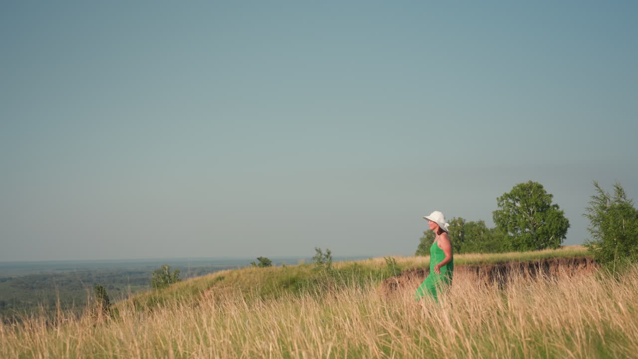 woman in green dress and white sunhat walks peacefully through tall dry grass beside edge of natural ditch, framed by trees and open field under clear blue sky on warm sunny day in countryside