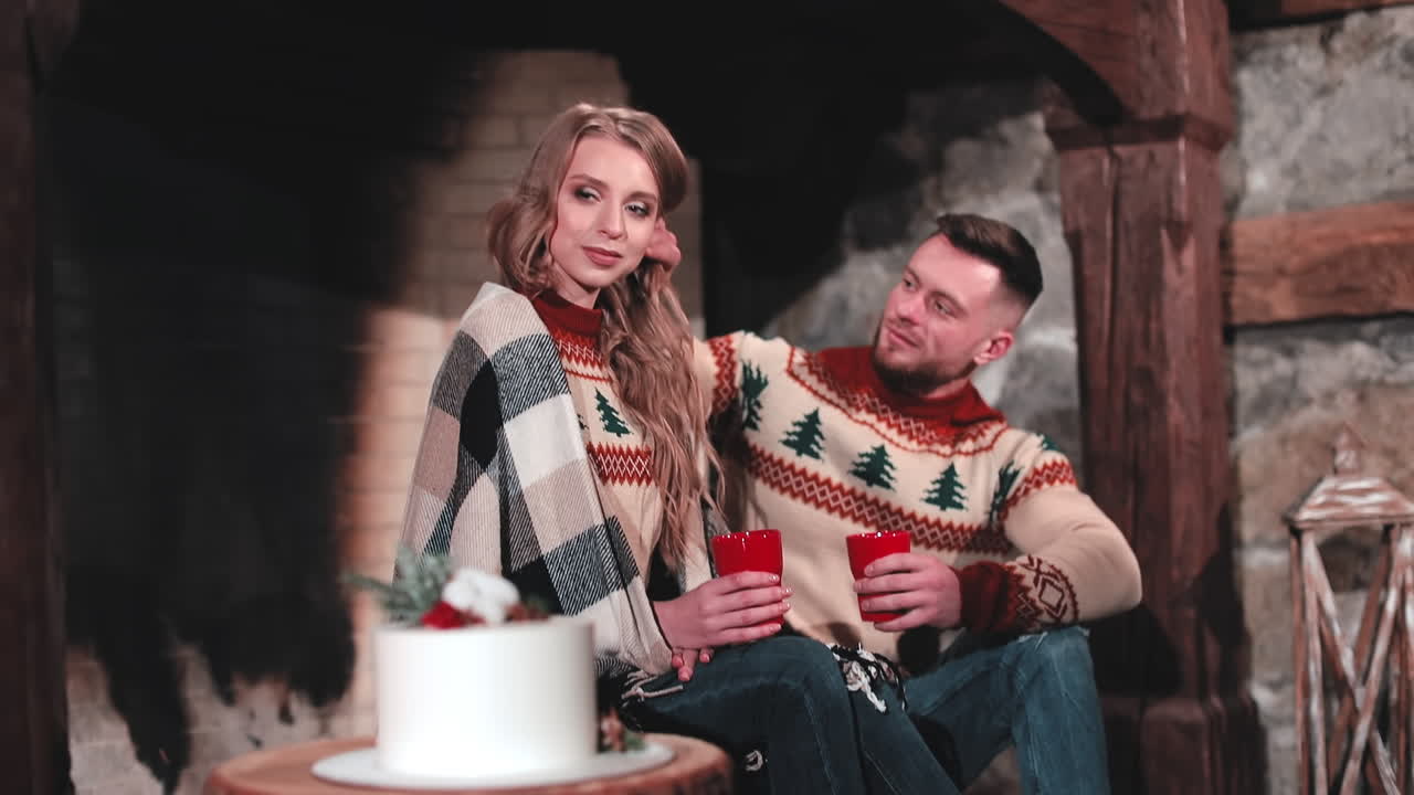 Beautiful young woman and her boyfriend near the fireplace. Couple wearing knitted Christmas sweaters sitting together in a country house.