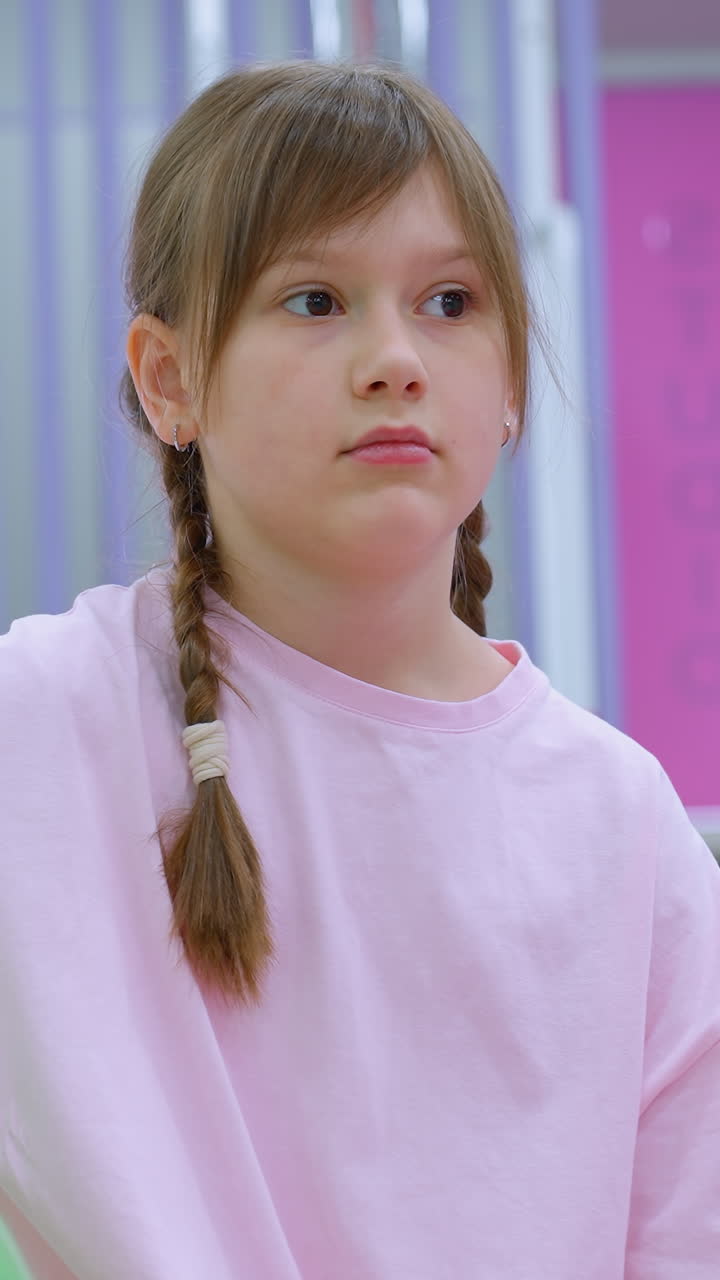 joven vestida de rosa se sienta cerca de la pelota de estabilidad verde en el gimnasio, apareciendo pensativa, el fondo presenta un moderno estudio de fitness con decoración púrpura y rosa, pisos suaves y equipos de ejercicio