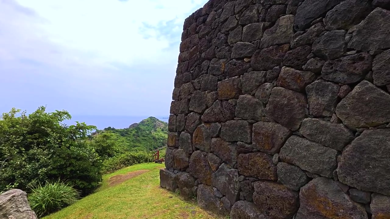 Primo piano di muratura in basalto eroso su muro curvo del faro a segnale di fumo Sanbang Yeondae, con promontorio costiero verde, mare e isola al largo sullo sfondo a Jeju, Corea del Sud