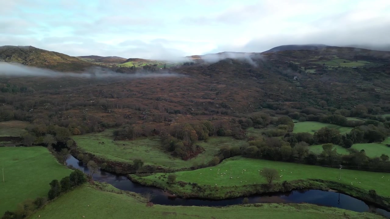 Virata aerea circolare sopra strada, prati, terreni agricoli, fiume con foresta e montagne nebbiose in lontananza. Contea di Kerry, Irlanda, in autunno.