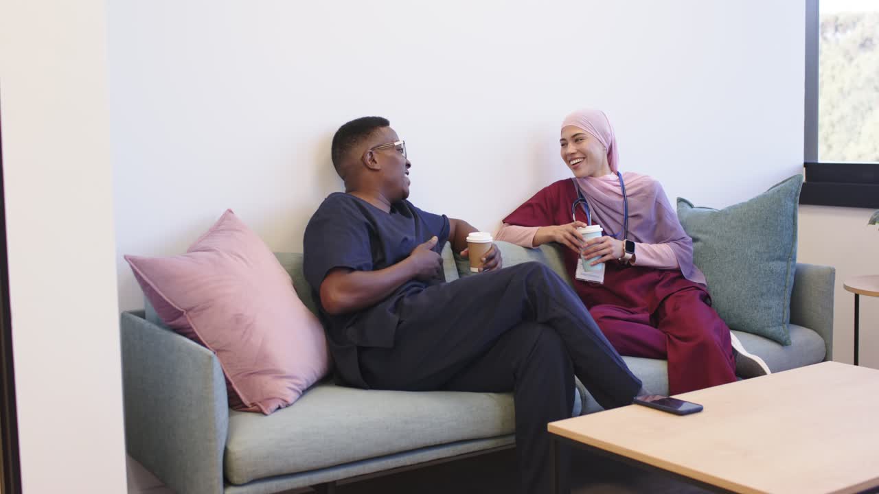 African American coworkers in scrubs sitting on couch on break, left opening cup lid, sharing laugh