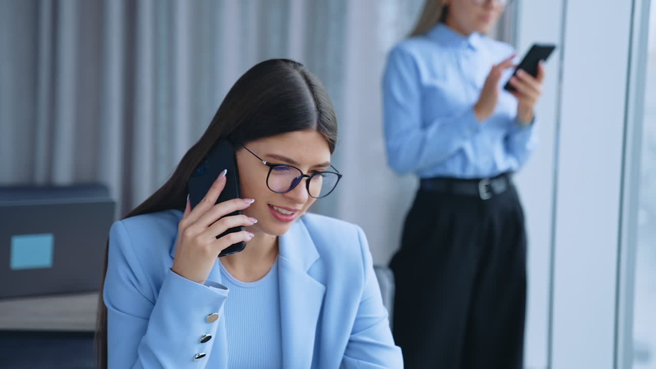 Smiling young beautiful female lady in glasses speaks on the phone sitting at desk. Blonde coworker stands at window messaging someone on smartphone.