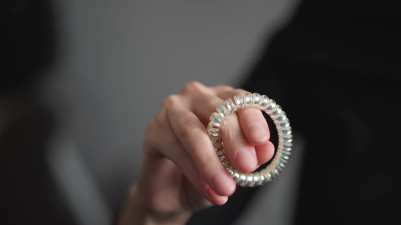 Primo piano di una mano che tiene e manipola un elastico per capelli a spirale iridescente su uno sfondo grigio tenue. La mano muove l'accessorio flessibile mostrandone la texture e le proprietà riflettenti alla luce naturale.