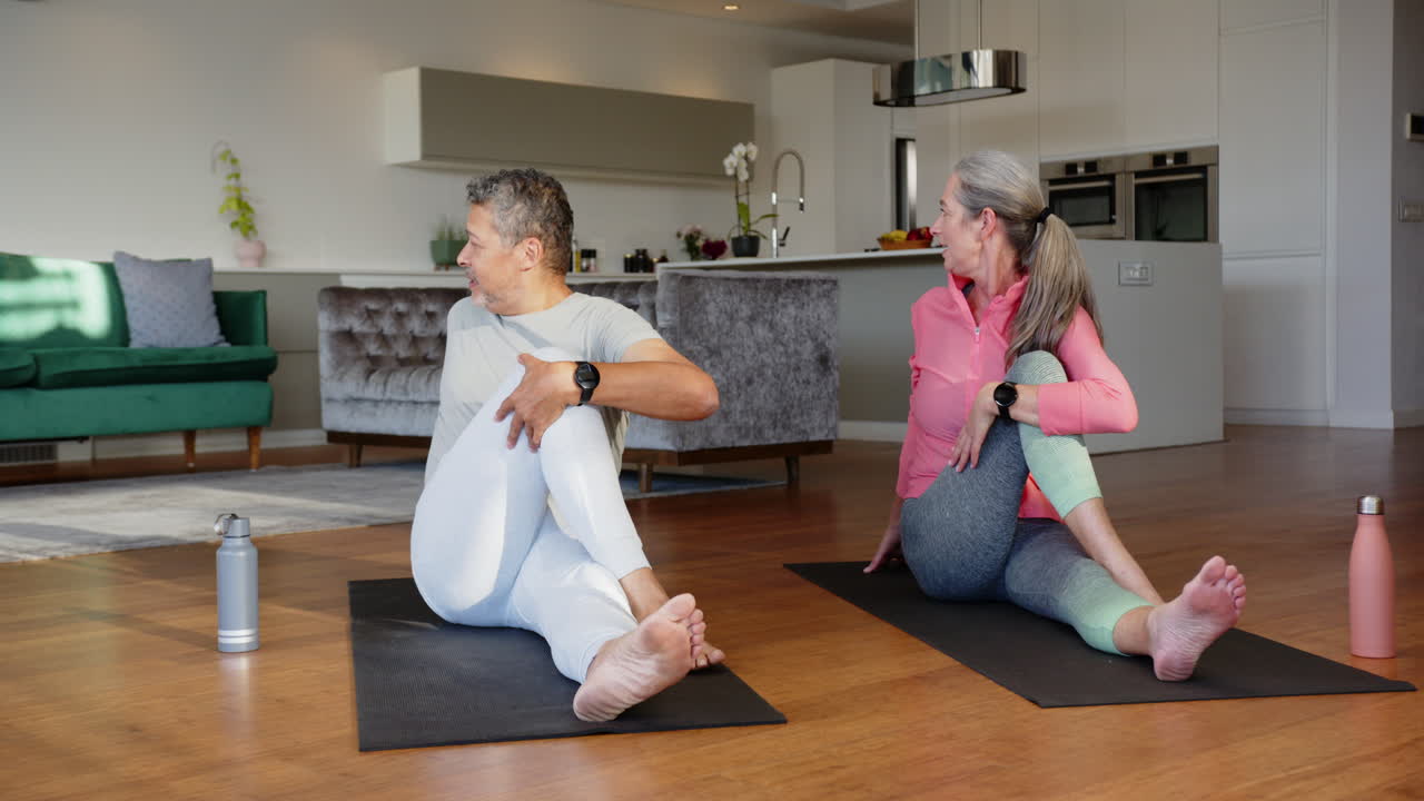 Practicing yoga, multiracial senior couple doing seated twist on yoga mats in living room, at home