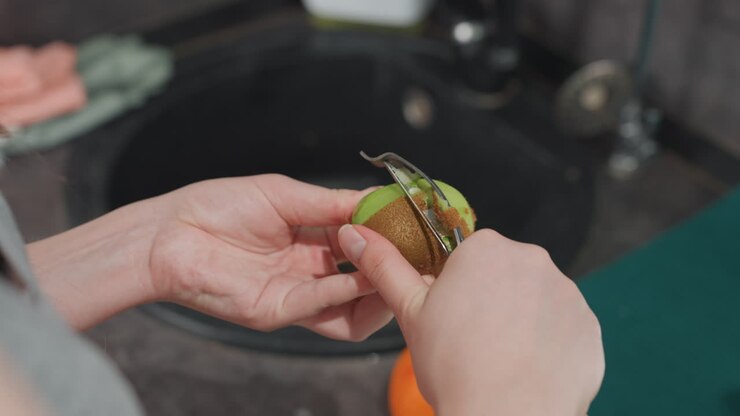 Woman with pregnancy peeling kiwi in kitchen. Expectant woman slices kiwi for healthy snack at home. Pregnant woman is preparing fresh kiwi snack using peeler at home in morning light