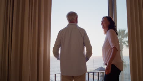 happy-old-travel-couple-arriving-in-hotel-room-on-vacation-looking-out-window-photographing-beautiful-view-of-ocean-at-sunset-enjoying-romantic-anniversary-holiday