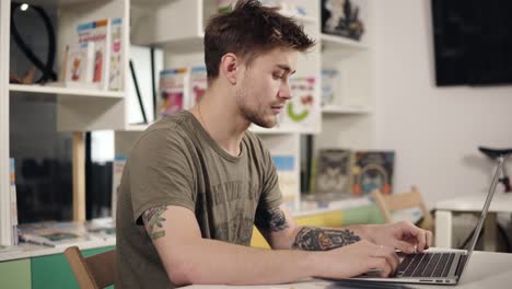 attractive young male programmer teacher with several tattooes on his arms is mumbling something while typing on laptop keyboard.