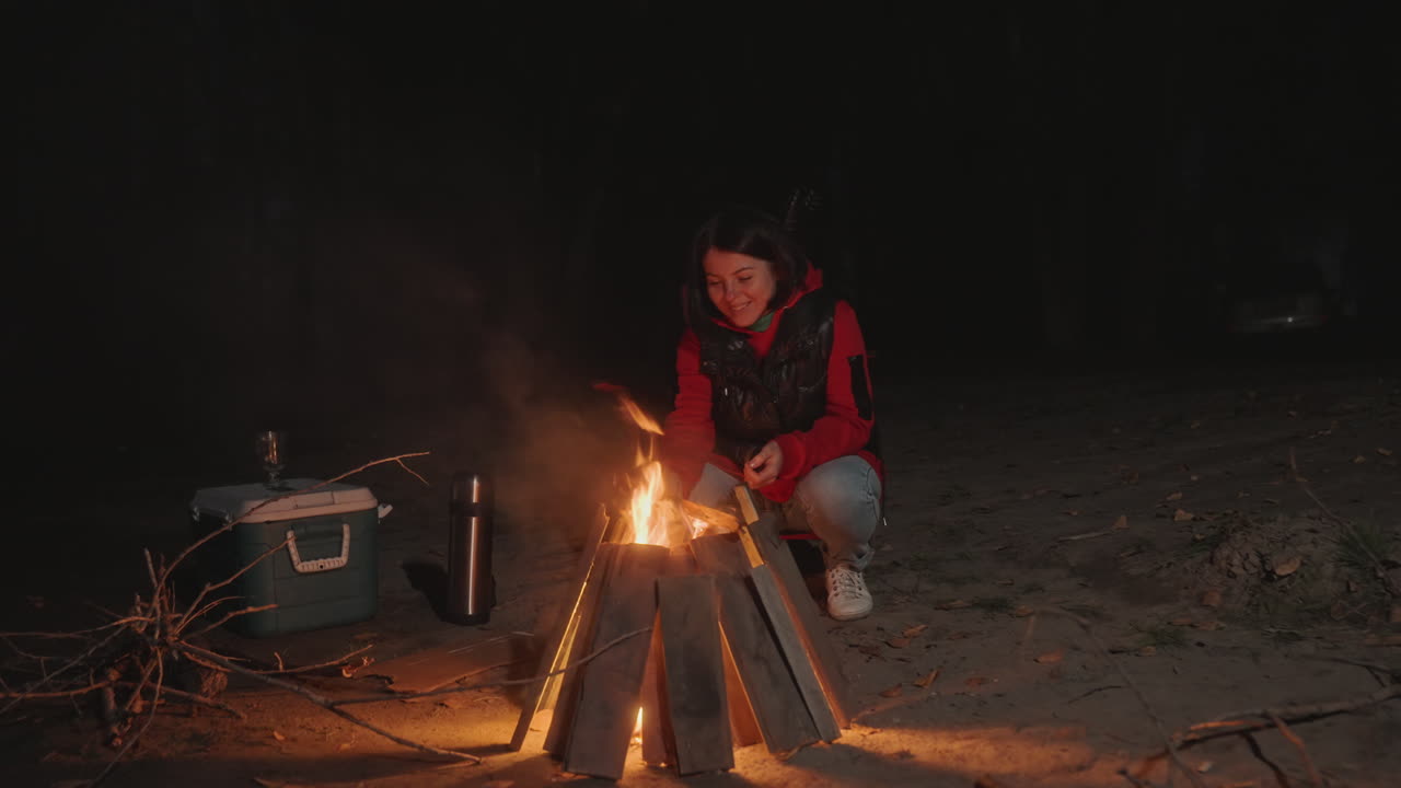 Mulher vestida de vermelho acende cuidadosamente uma fogueira aconchegante sob o céu noturno.