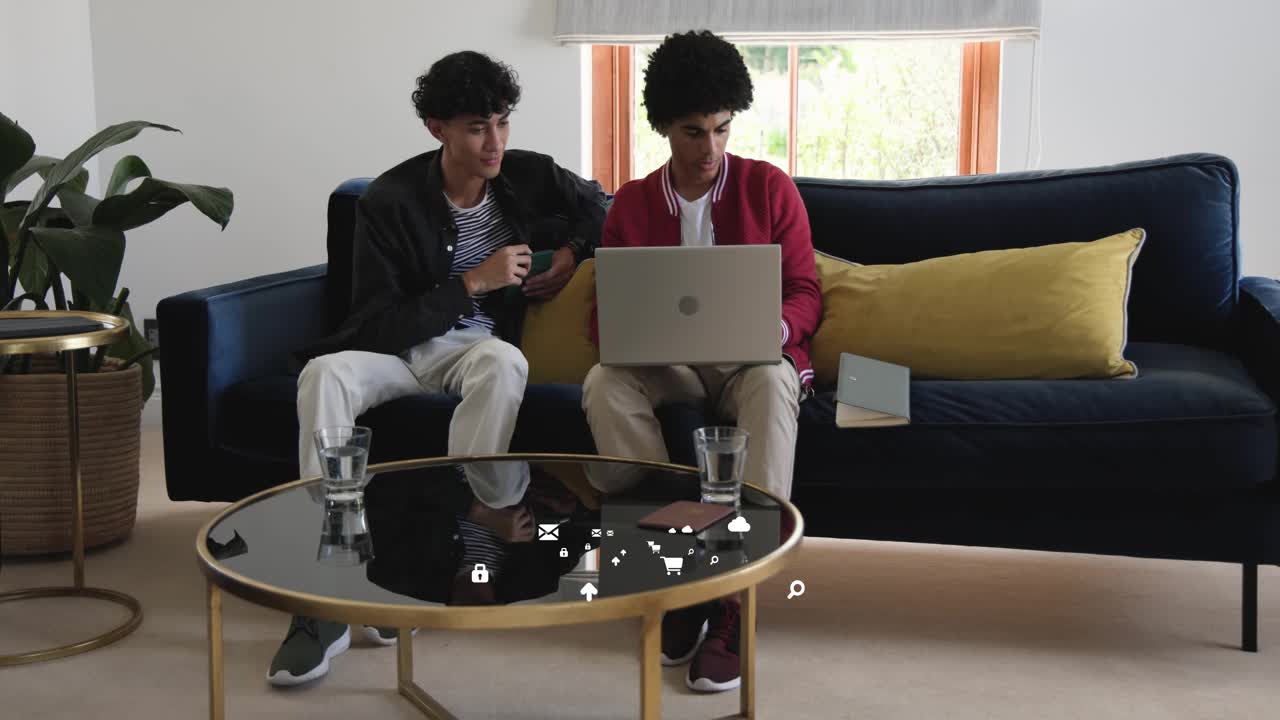 Two men leaning on sofa, one pointing device causing floating app icons while collaborating on tech