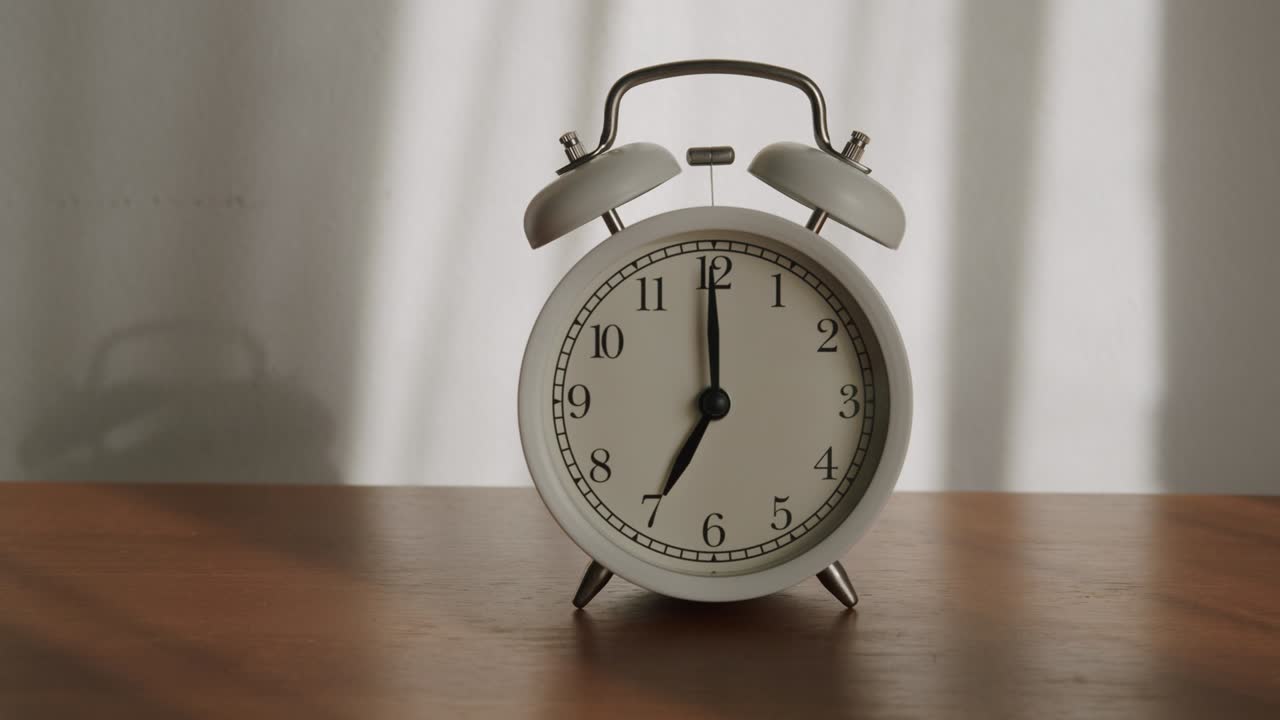 White alarm clock is placed on a wooden table and the light shines through the curtains in the morning.