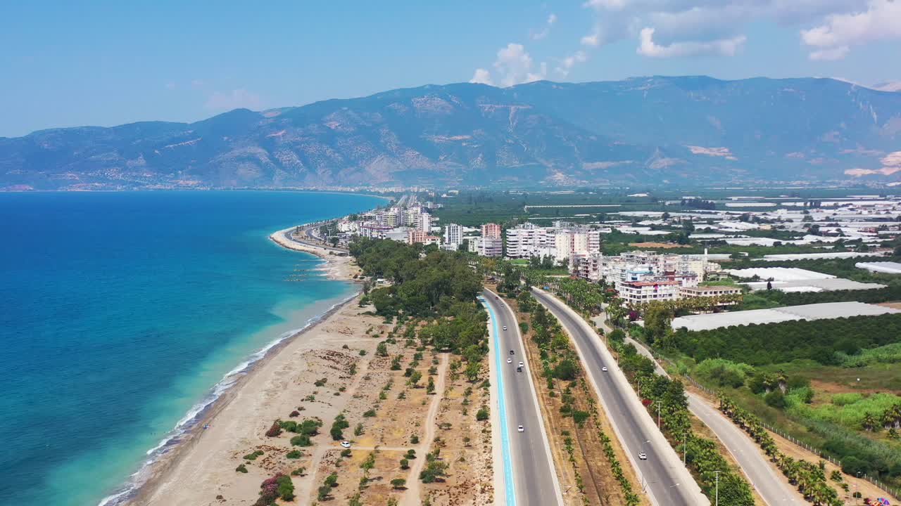 Wide aerial view of an empty highway road leading a coastal beach town with tropical blue Mediterranean Sea and large mountains in the distance