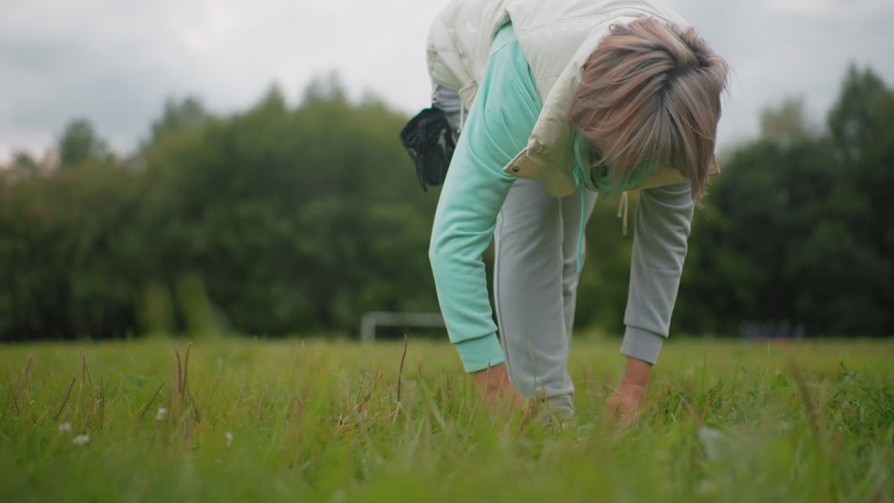Dog instructor wearing mint green hoodie and light puffy vest bends down on grassy field adjusting canvas shoe after training session, focused on comfort and readiness in calm outdoor environment