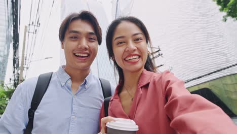 two smiling young adults taking a selfie with coffee on a city street