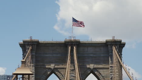 the us flag waving in the wind on top of the brooklyn bridge on a sunny day with a blue sky and some clouds, static shot