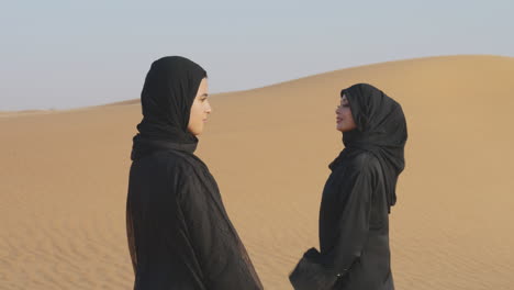 due belle donne musulmane in hijab in piedi in un deserto ventoso e sorridono alla macchina fotografica