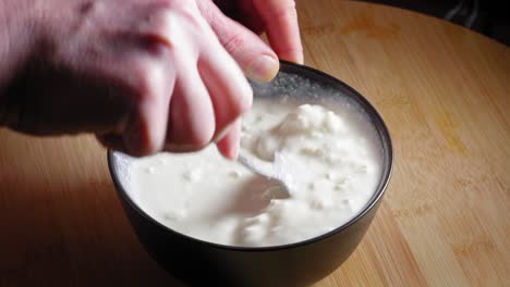 a close up shot of a black mixing bowl on a wooden kitchen table filled with white low fat cheese as a chef carefully breaks up clumps with the back of a spoon and stirring to create an even texture