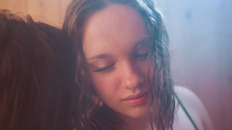 close up of sisters leaning together in warm wood paneled interior, one resting head on sibling back while soft backlight casts gentle haze over wet hair and closed eyes