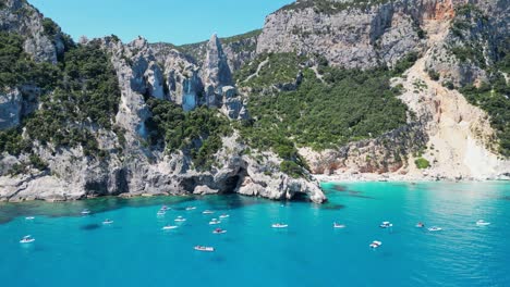 tourist boats relax at summer vacation in cala goloritze beach, sardinia, italy - aerial 4k circling