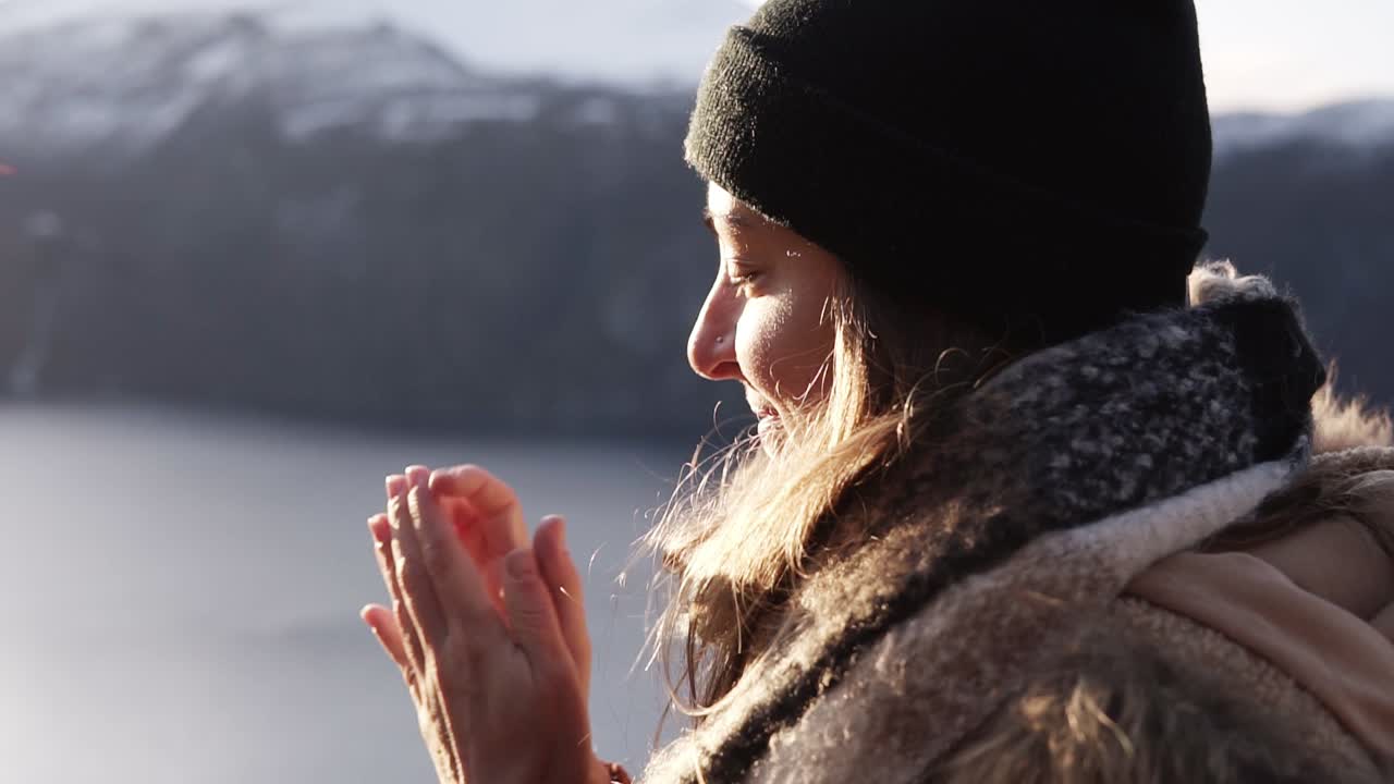 mujer sonriente en primer plano de abrigo de invierno en un día de invierno, retrato mirando directamente al horizonte con un paisaje increíble de fondo con lago y montaña nevada. mujer admirando la vista, calentándose las manos debido al clima frío.