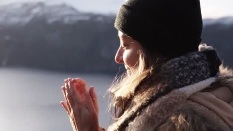 smiling woman in winter coat closeup in a winter day , portrait looking straight to the horizon with background amazing landscape with lake and snowy mountain. woman admiring the view, warming her hands because of a cold weather