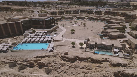 Drone-flies-over-a-beautiful-architecture-stone-built-beresheet-hotel-in-a-desert-landscape,-Flying-back-and-reveals-the-beautiful-cliff-1