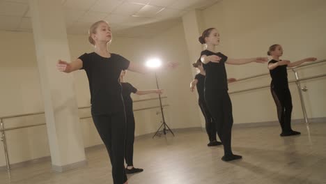 a group of young ballet students in black dancewear practicing positions in a spacious ballet studio with wooden flooring and wall-mounted barres. focused expressions and synchronized movements.