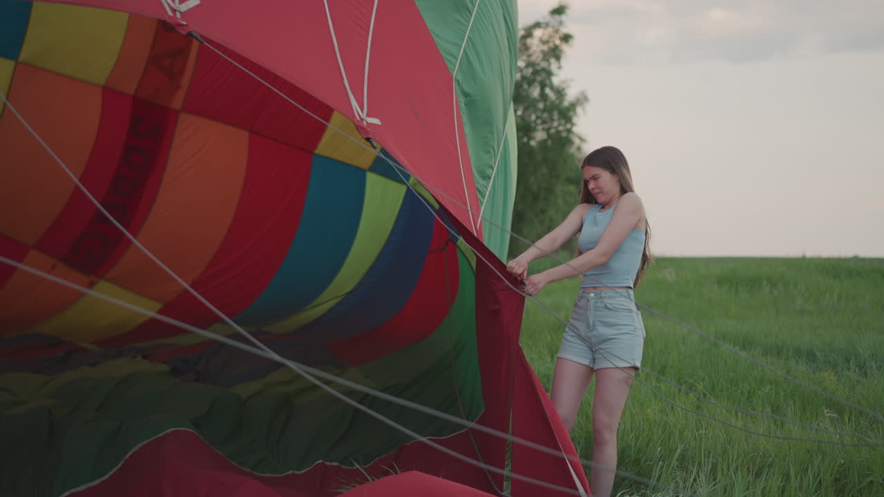 woman grips hot air balloon envelope near roaring burner flame during preflight setup in lush green field under soft pastel sky with gentle breeze lightly stirring colorful fabric panels