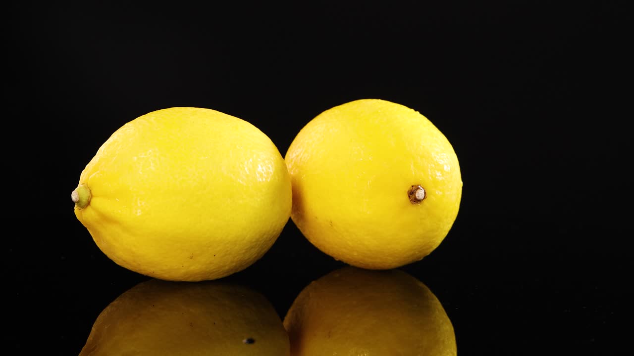 Two fresh lemons rotate together on a reflective black surface under bright studio lighting, creating dynamic reflections and a clean, minimal aesthetic