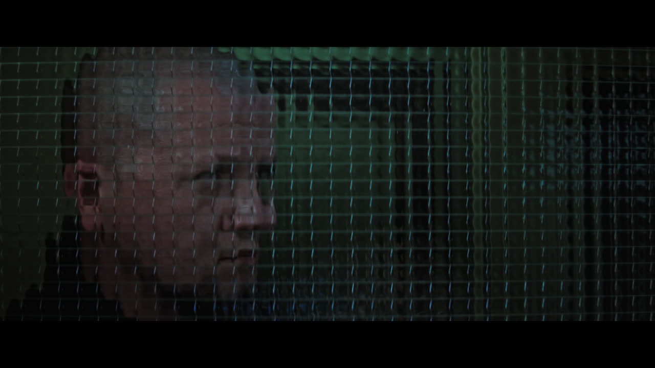 A bald dangerous looking man checks the corridor through reinforced glass