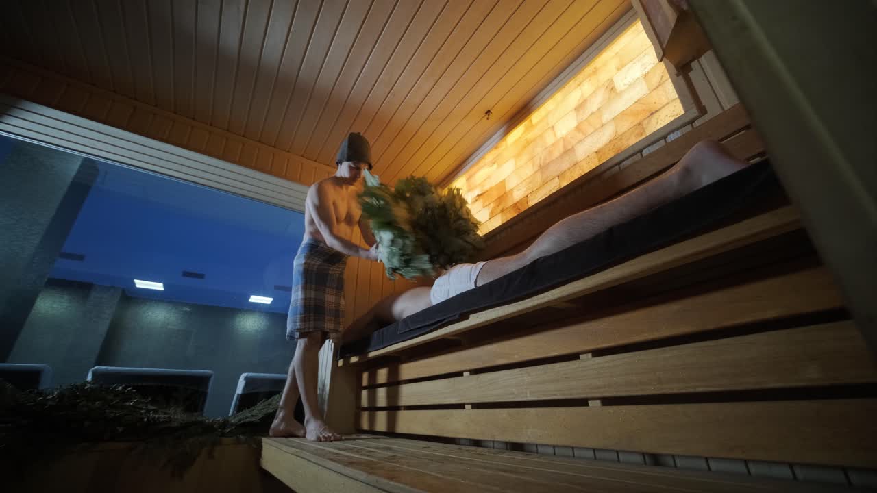 hombre tendido en un banco de madera en el spa con masajista haciendo masaje con escoba de roble. cliente en sauna finlandesa en el interior con profesional.