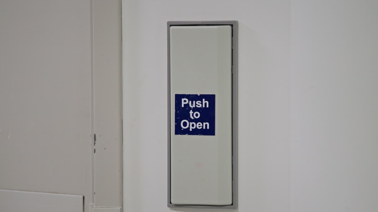 Close-up of a hand pressing a “Push to Open” button on a wall-mounted door release panel in a hospital. The image highlights accessibility features in healthcare environments