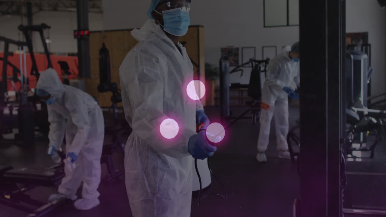 People in protective suits disinfecting gym equipment with glowing animation effects