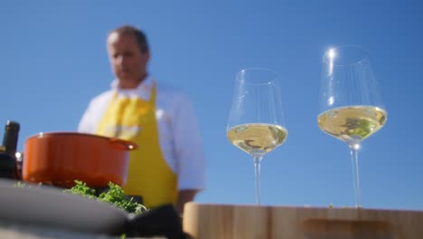 dos copas de vino blanco en primer plano mientras un cocinero está de pie en el fondo con un delantal de cocina amarillo