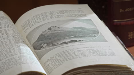 Close-up-of-an-open-book-with-a-text-and-a-drawing-of-a-castle-on-the-mountain,-Next-to-the-open-book-is-an-closed-vertivally-put-big-book