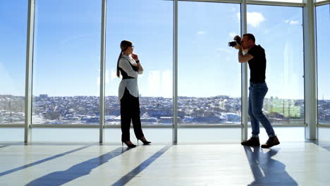 attractive business lady posing to the photographer. pretty young woman in glasses and elegant clothes standing near the large office window. man taking photos of a business woman in light office. profile view.